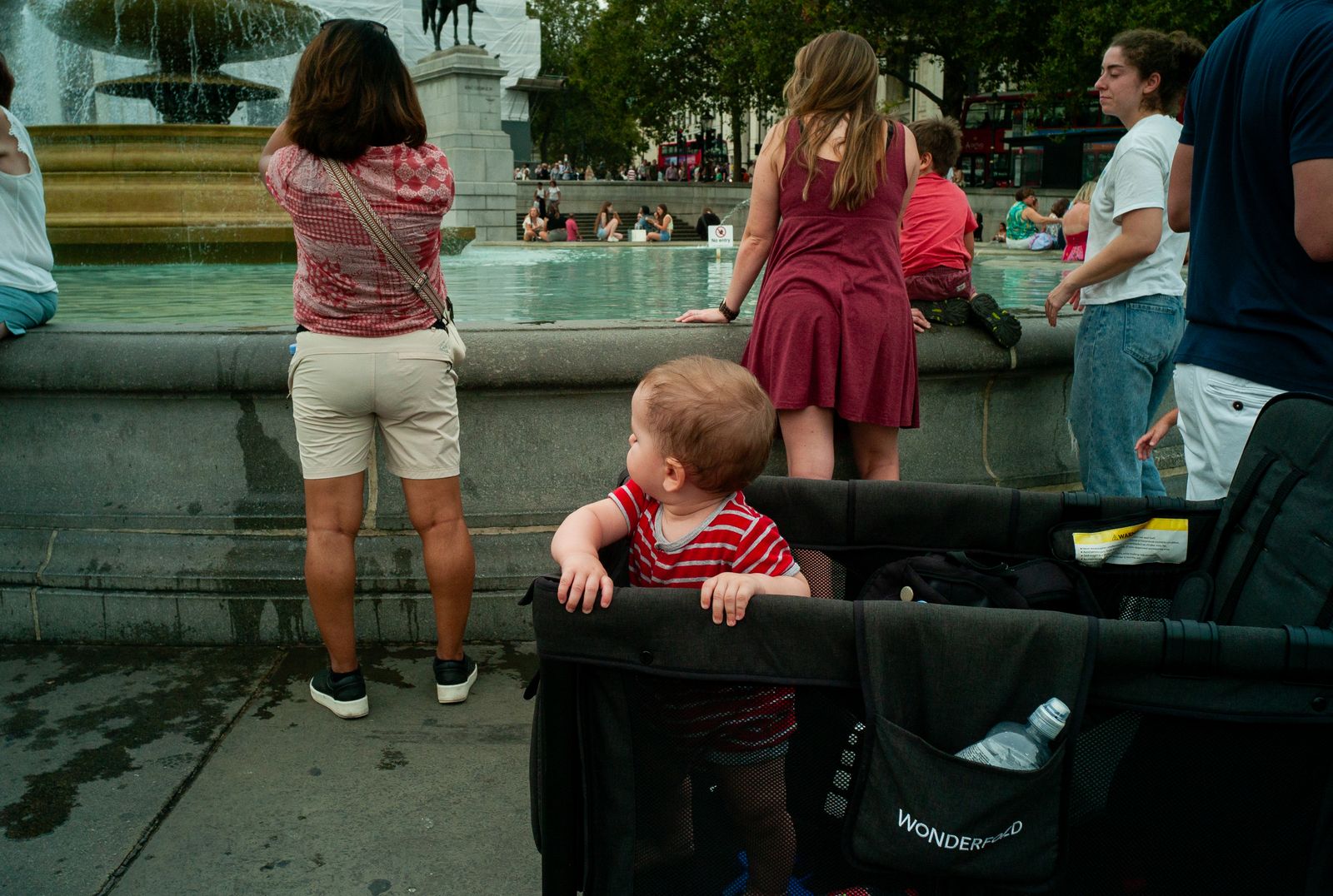 a little boy sitting in a stroller in front of a fountain