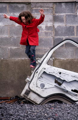 a girl in a red jacket jumping over a broken car