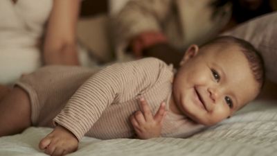 a smiling baby laying on a bed with a woman in the background