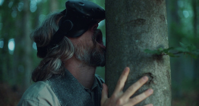 a man with long hair and a beard wearing headphones leans against a tree trunk