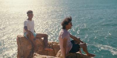 two young men sitting on a cliff overlooking the ocean