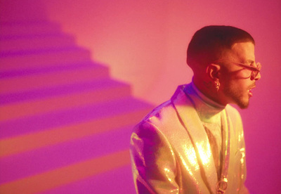 a man in a gold jacket and glasses standing in front of a pink background