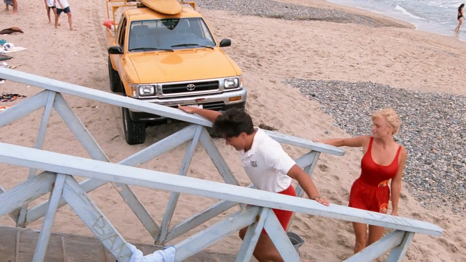 a man and a woman walking up a beach next to a yellow truck