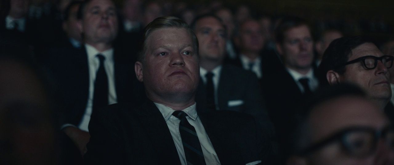 a man in a suit and tie sitting in front of a crowd