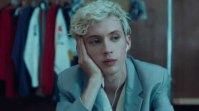 a man with blonde hair sitting in front of a rack of clothes