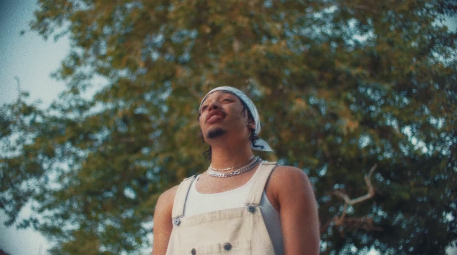 a man in overalls and a headband standing in front of a tree