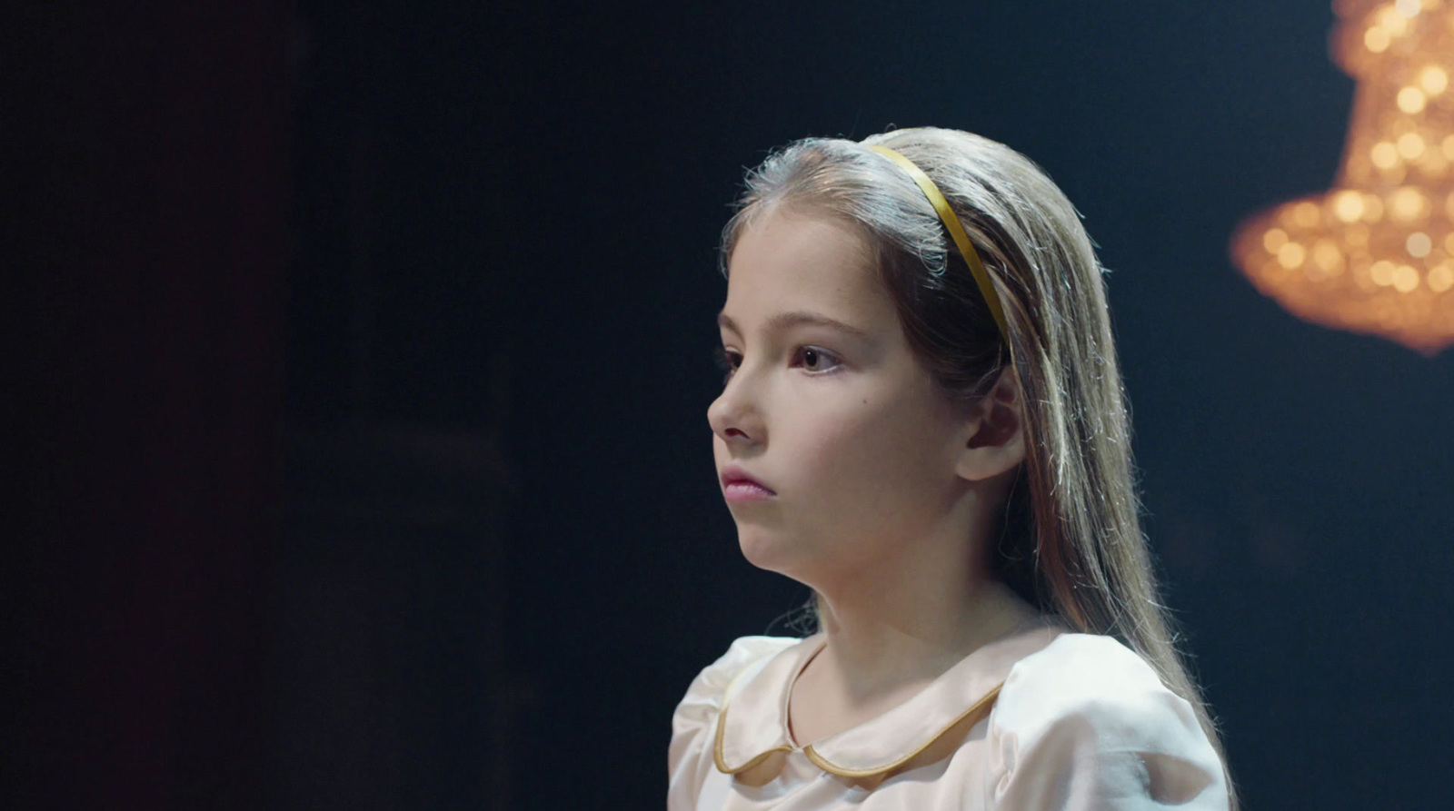 a little girl with a ponytail standing in front of a chandelier